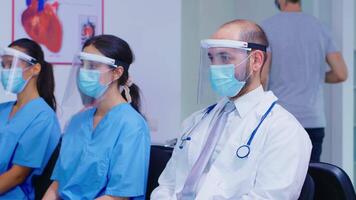 grupo do médico funcionários sentado dentro hospital esperando área vestindo face mascarar e viseira contra coronavírus. médico com branco casaco e estetoscópio. paciente deixando clínica. video