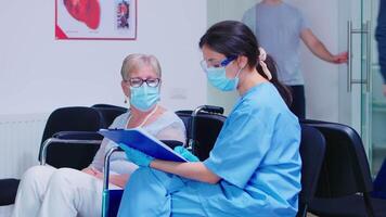 Nurse explaining diagnosis to disabled senior woman in wheelchair. Assistant wearing face mask against infection with coronavirus in hospital waiting area. video