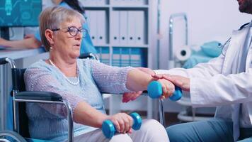 Physiotherapy for elderly woman in wheelchair, she is practicing with dumbbells to recover after a musle trauma. Modern private hospital or rehabilitation clinic. Lifting exercise for disabled people video