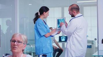 Medical staff holding tablet pc with patient radiography in hospital hallway. Doctor discussing diagnosis with nurse in clinic waiting area. Old woman sitting in wheelchair. video