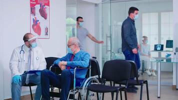 inválido velho homem com face mascarar contra infecção com coronavírus dentro cadeira de rodas discutindo com médico dentro hospital esperando área. enfermagem convidativo paciente dentro exame sala. video