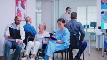 Doctor with stethoscope holding disabled senior woman radiography in wheelchair while talking with her in hospital waiting area. Patient asking about his appointment at clinic reception. video