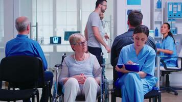 Nurse filing documents while talking with disabled senior woman in hospital waiting area. Patient asking for direction at hospital reception. video