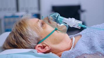 Close up portrait of retired woman breathing with difficulties in oxygen mask lying in hospital bed during covid-19 coronavirus outbreak. Medical medicine healthcare system. Infection treatment video