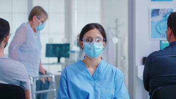 femmina infermiera con viso maschera contro coronavirus nel ospedale in attesa la zona guardare a telecamera. anziano donna con a piedi invalidità utilizzando zimmer telaio. video