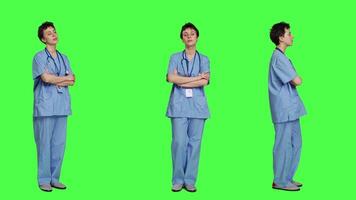 retrato do sorridente médico assistente poses com braços cruzado, mostrando confiança vestido dentro azul hospital esfrega. bem sucedido enfermeira em pé contra tela verde pano de fundo, saúde especialista. Câmera uma. video