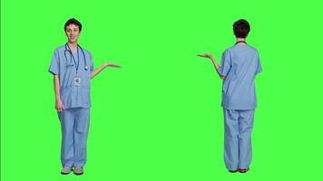Medical assistant pointing at something aside and doing web commercial, creating advertisement against greenscreen backdrop. Young nurse with scrubs showing a thing to left or right sides. Camera A. video