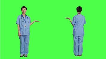 Medical assistant pointing at something aside and doing web commercial, creating advertisement against greenscreen backdrop. Young nurse with scrubs showing a thing to left or right sides. Camera A. video