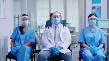 Team of medical staff with protection mask and visor against coronavirus in hospital waiting area looking at camera. Patient in examination room for consultation. video