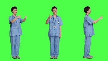 Cheerful nurse giving thumbs up sign against greenscreen backdrop, showing positive gesture with a like. Medical assistant shows her approval, recommending something and being satisfied. Camera A. video