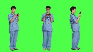 Medical assistant playing mobile video games on smartphone app, taking a break from healthcare work. Nurse enjoys gaming tournament on phone, standing against greenscreen backdrop. Camera A.