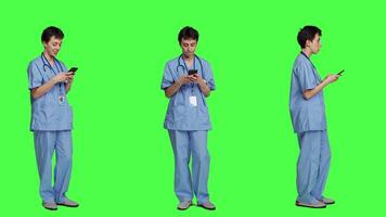 Young nurse texting on smartphone against greenscreen backdrop, using mobile phone to check social media messages on work break. Medical assistant browsing online web pages. Camera A. video