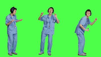 Nurse having fun listening to music on wireless headphones, wearing scrubs and dancing around against greenscreen backdrop. Medical assistant showing groovy funky dance moves. Camera A. video