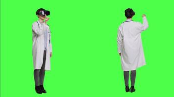 Woman medic works with 3d virtual reality headset in studio, standing against greenscreen backdrop. General practitioner uses vr glasses with interactive vision tool, futuristic exam. Camera A. video