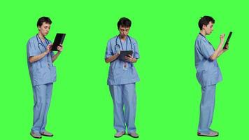 Medical assistant browsing online webpages on tablet and texting, using social media apps to chat with people. Nurse navigates internet on gadget, standing against greenscreen backdrop. Camera A. video