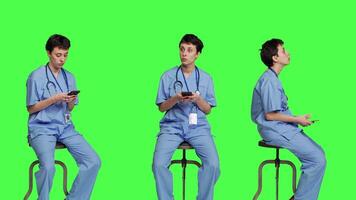 Nurse browsing online websites on smartphone app in studio, sitting on a chair against greenscreen backdrop. Medical specialist in scrubs using mobile phone and social media texting. Camera A. video