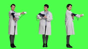 Healthcare specialist using a tonometer to measure high blood pressure and pulse, presenting tool used at checkup examinations. Doctor in white coat stands against greenscreen backdrop. Camera A. video