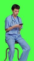 Side view Nurse browsing online websites on smartphone app in studio, sitting on a chair against greenscreen backdrop. Medical specialist in scrubs using mobile phone and social media texting. Camera A. video