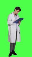 Side view Physician taking notes after patient examination against greenscreen backdrop, writing down medicaments to cure illness. Doctor in white coat using clipboard papers for checkup. Camera A. video