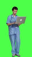 Front view Medical assistant browsing for healthcare treatments to help patients, using laptop against greenscreen backdrop. Young nurse navigates online websites an searches for health support. Camera A. video