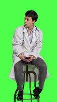 Front view Woman specialist using smartphone social media apps in studio, sitting on a chair against greenscreen backdrop. Physician waiting for someone and browses online webpages, texting. Camera A. video