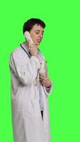 Side view Physician answering landline phone call against greenscreen backdrop, talking to patients about checkup appointments. Medic having remote discussion on retro telephone with cord. Camera B. video