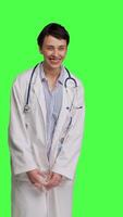 Front view Portrait of general practitioner laughing at something against greenscreen backdrop, feeling joyful and confident with her healthcare expertise. Woman medic in white coat smiling in studio. Camera B. video