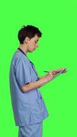 Profile Medical assistant writing checkup information on clipboard files, taking notes and making doctor appointments with checklist. Nurse wearing blue scrubs stands against greenscreen backdrop. Camera B. video