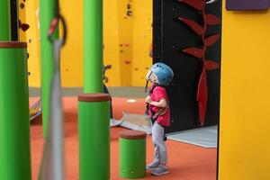 niño alpinismo en pared en diversión centro. alpinismo formación para niños. pequeño niña en vestido alpinismo engranaje escalada alto. extremo activo ocio para niños. foto