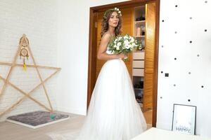 The bride in a beautiful wedding dress with a long curly hair stands in the bedroom and looks at the camera. Bride indoors with white flowers bouquet and tender wreath hairstyle photo