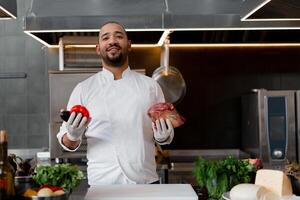 contento sonriente cocinero prepara carne plato con varios vegetales en el cocina. en uno mano el hombre sostiene verduras, en el otro un pedazo de Fresco carne foto