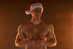 Portrait of muscular man wearing Christmas Santa hat, showing thumb up on smoky background photo