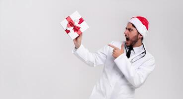 Young handsome doctor in white uniforme and Santa Claus hat standing in studio on white background smile and Shows a finger at the gift box. photo