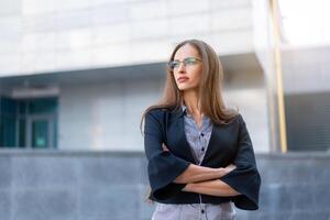 Business woman with long hair dressed black jacket standing outdoor near corporate office building hands folded photo