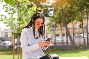 Young woman hipster with dreadlocks in pink sunglasses use smartphone sitting summer park outdoor. New generation, Technology in hand. photo