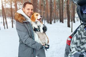 A young man dressed in a gray winter park in a snowy winter forest holds a dog named Beagle. Concept about winter leisure and love to animals photo