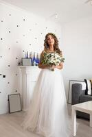 The bride in a beautiful wedding dress with a long curly hair stands in the bedroom and looks at the camera. Bride indoors with white flowers bouquet and tender wreath hairstyle photo