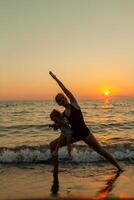 woman and girl silhouette practicing balancing yoga warrior pose together during ocean sunset with bright orange sky and water reflections photo