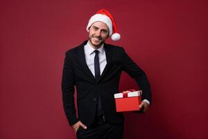 joven hermoso caucásico chico en negocio traje y Papa Noel sombreros soportes en rojo antecedentes en estudio y dientes carita participación rojo regalo caja en mano. foto