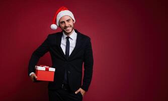 joven hermoso caucásico chico en negocio traje y Papa Noel sombreros soportes en rojo antecedentes en estudio y carita participación rojo regalo caja en mano. foto