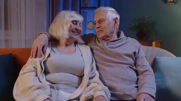 Happy old senior elderly family couple hugging, laughing, smiling looking at camera at home sofa video