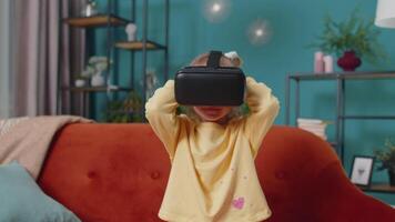 Toddler girl sitting on home sofa using virtual reality headset helmet app to play video 3D game