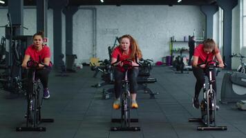 grupo de sonriente amigos mujer clase hacer ejercicio, capacitación, hilado en estacionario bicicleta a moderno gimnasio video