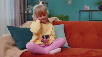 Child girl kid with smartphone in headphones dancing while listening music at home alone on sofa video