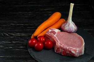 Top view of one pieces raw pork chop steaks with cherry tomatoes carrot and garlic on a black stone cutting board. photo