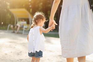 pequeño caucásico niña participación su madre por el mano para un caminar en un verano parque. fuerte familia y amor de niños y padres. foto