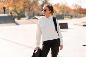 Outdoor portrait of young beautiful woman with a ponytail and sunglasses, with a backpack on his shoulders, dressed in a white sweater, near the sportsground photo