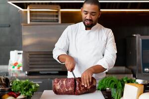 Handsome young African chef standing in professional kitchen in restaurant preparing a meal of meat and cheese vegetables. photo