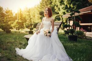 Bride in elegant wedding gown sits on stone bench photo