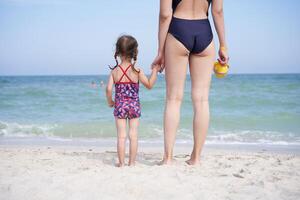 madre hija playa juntos posterior ver irreconocible caucásico mujer pequeño niña trajes de baño en pie playa atrás. foto